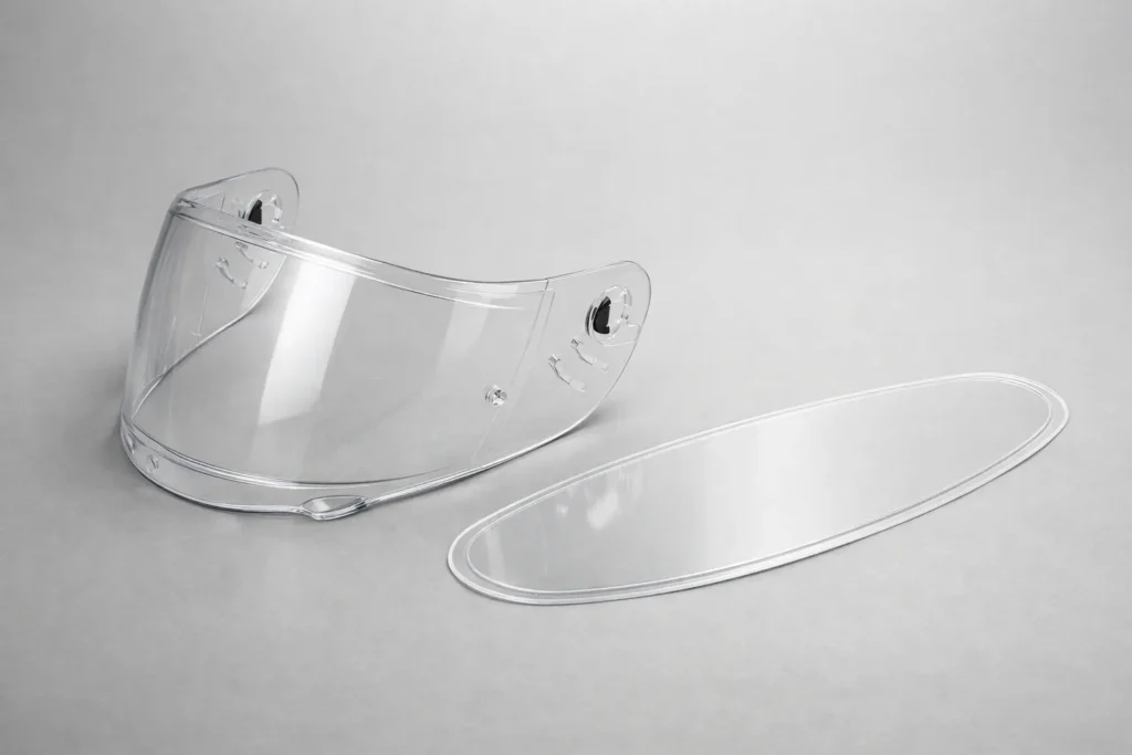 Une visière de casque moto transparente et un insert antibuée amovible posés côte à côte.